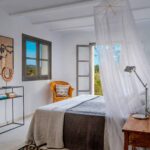 Bedroom with white mosquito net over the bed, orange wicker armchair, zigzag rug and window onto the landscape