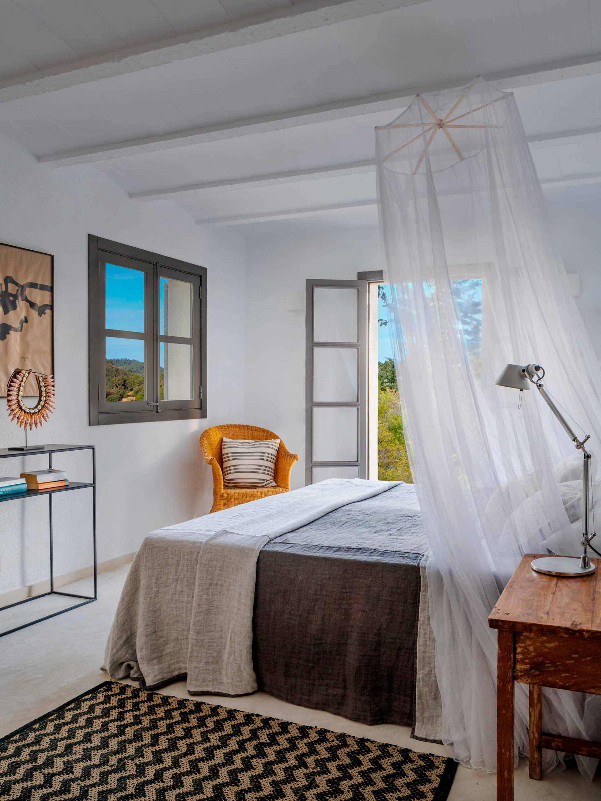 Bedroom with white mosquito net over the bed, orange wicker armchair, zigzag rug and window onto the landscape