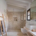 Bathroom with white beams, shower clad in pebble mosaic, pale stone floor and countertop basin