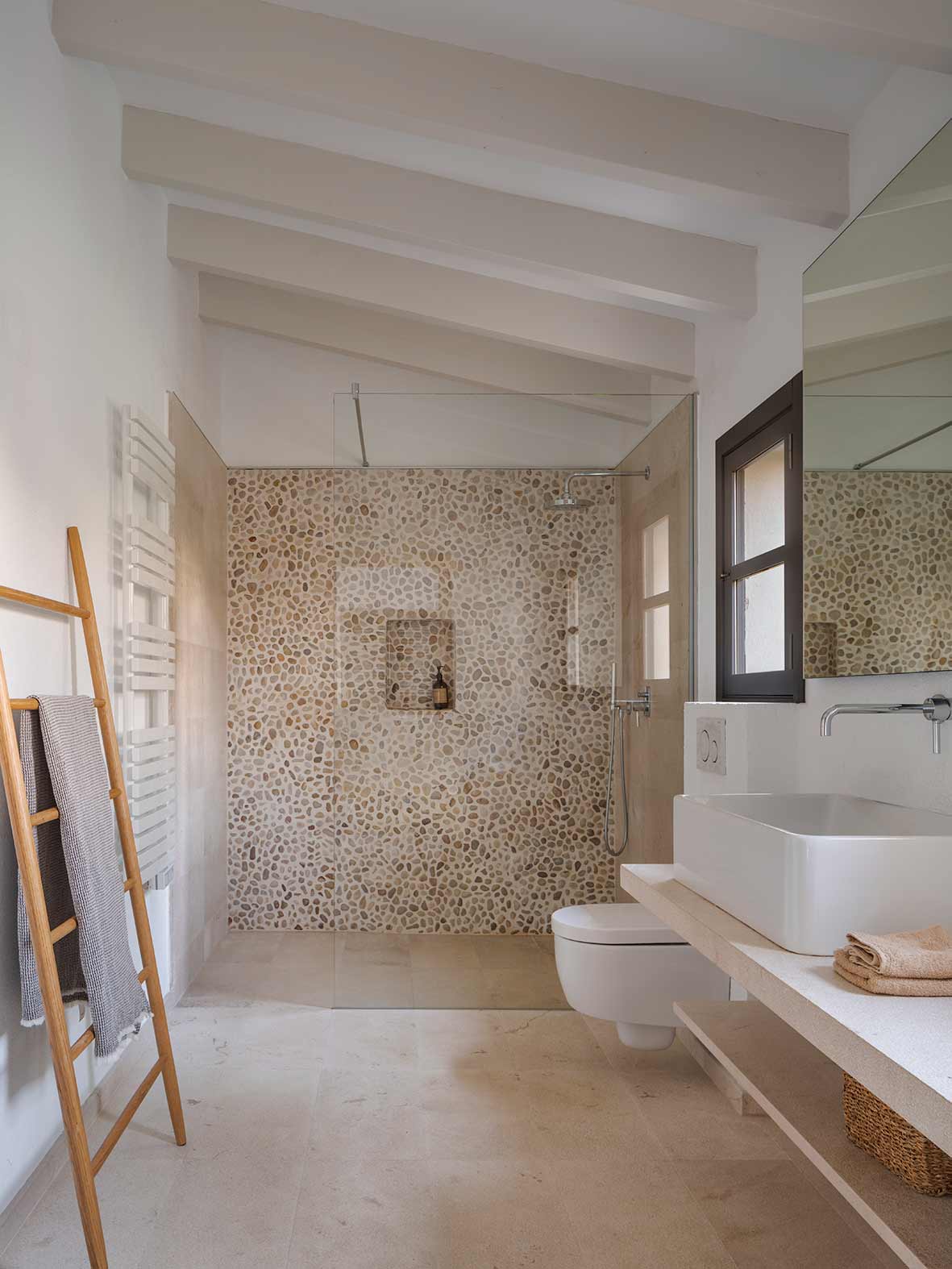 Bathroom with white beams, shower clad in pebble mosaic, pale stone floor and countertop basin