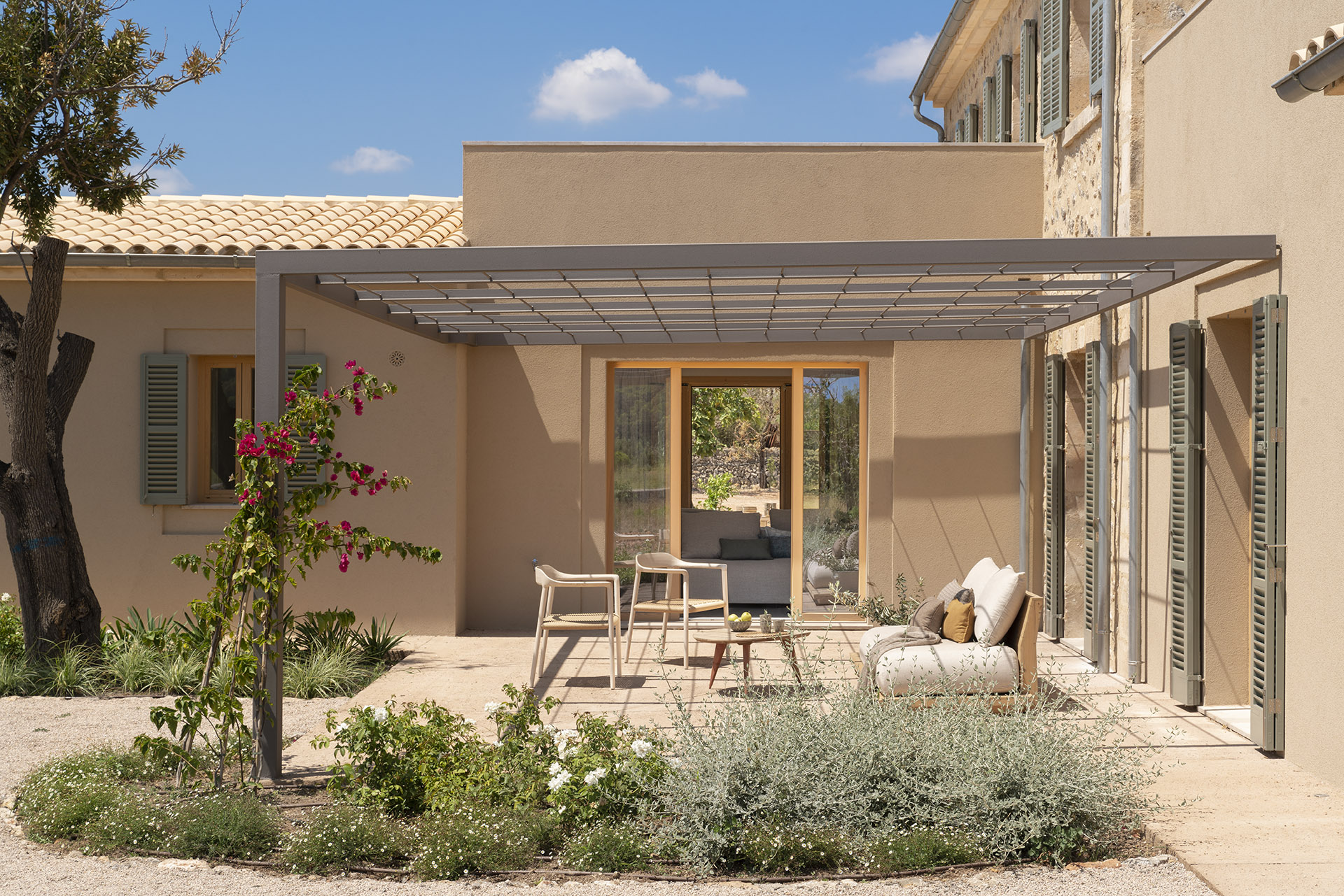 Terrasse mit Metallpergola, Außensofa und hellen Stühlen zwischen Pflanzen, neben der Natursteinfassade
