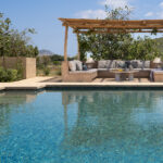 Swimmingpool mit türkisfarbenem Wasser, Ruhebereich unter einer Holzpergola und durchgehenden Sitzbänken im Hintergrund