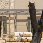 Außensofa mit ockerfarbenen Kissen auf Holzdielen, neben einem Baumstamm und einer Metallpergola