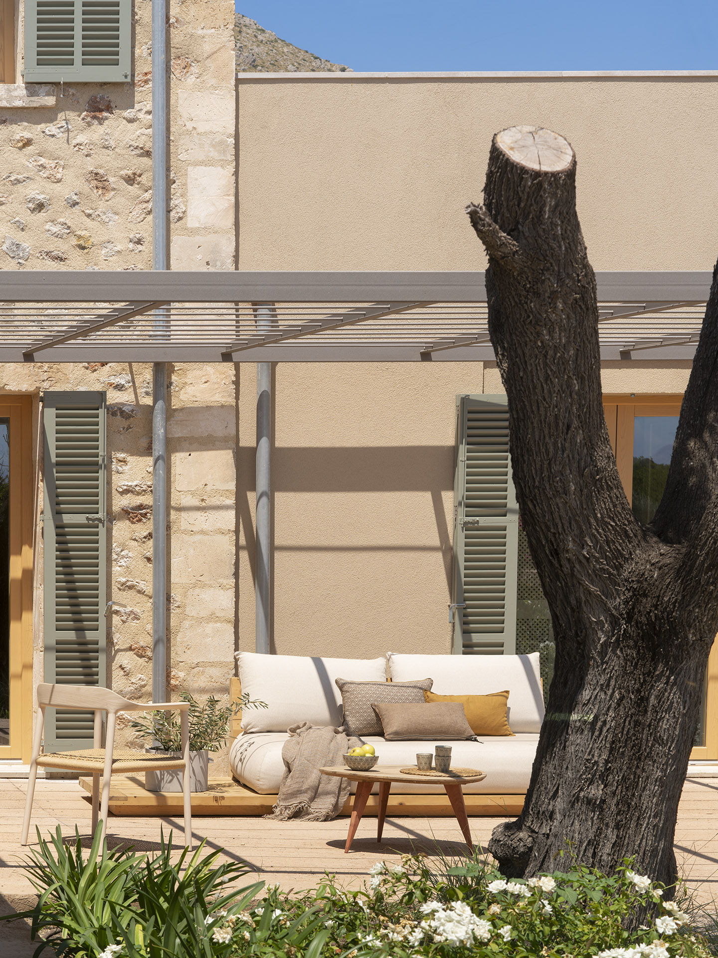 Außensofa mit ockerfarbenen Kissen auf Holzdielen, neben einem Baumstamm und einer Metallpergola