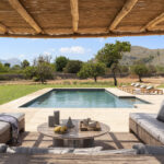 Chill-out-Bereich unter einer Schilfpergola mit durchgehenden Sofas, rundem Tisch und Blick auf den Swimmingpool