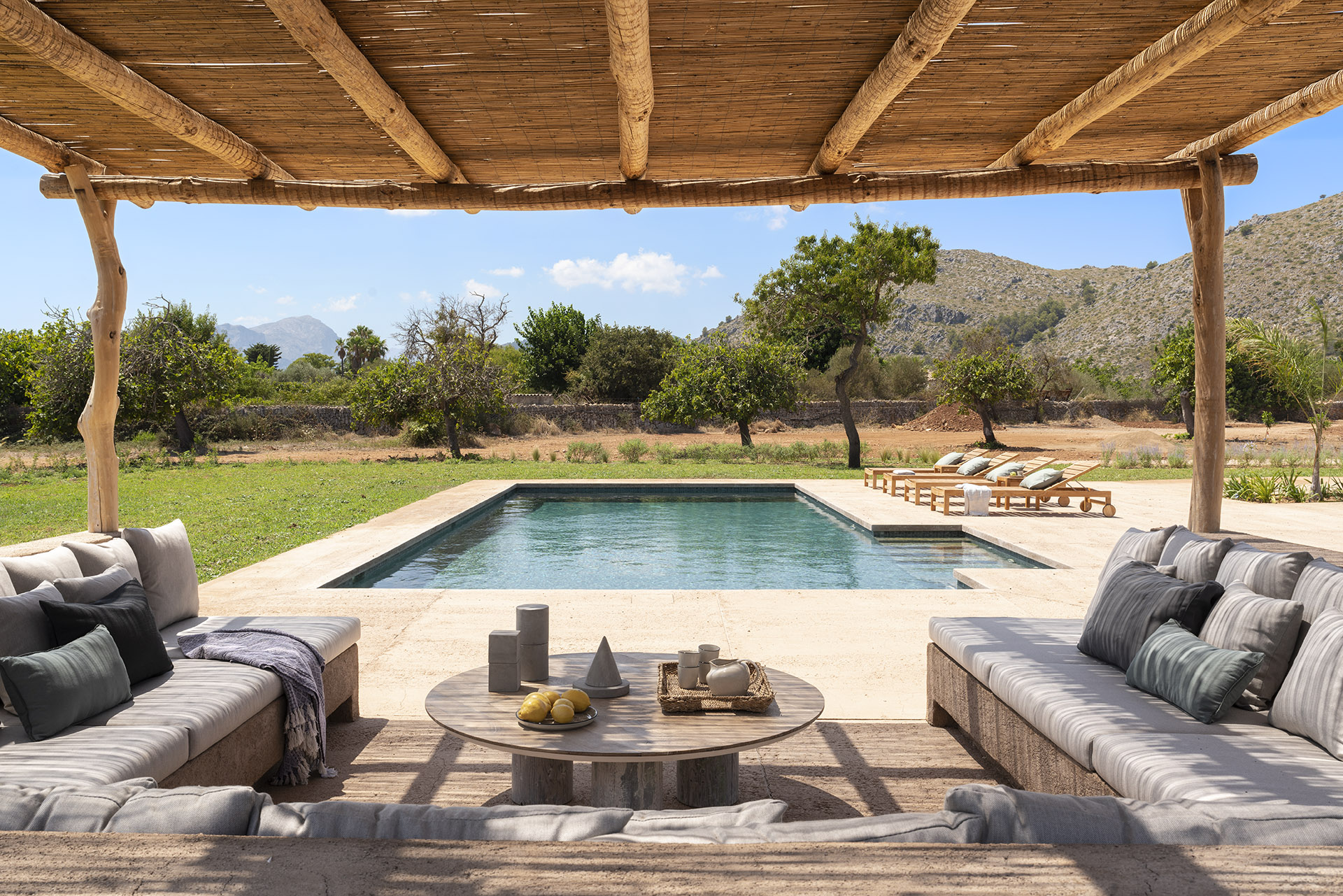 Chill-out-Bereich unter einer Schilfpergola mit durchgehenden Sofas, rundem Tisch und Blick auf den Swimmingpool