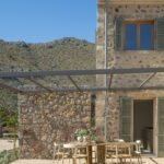 Steinfassade mit grünen Fensterläden, Metallpergola und Außentisch mit hellen Stühlen vor der Bergkulisse