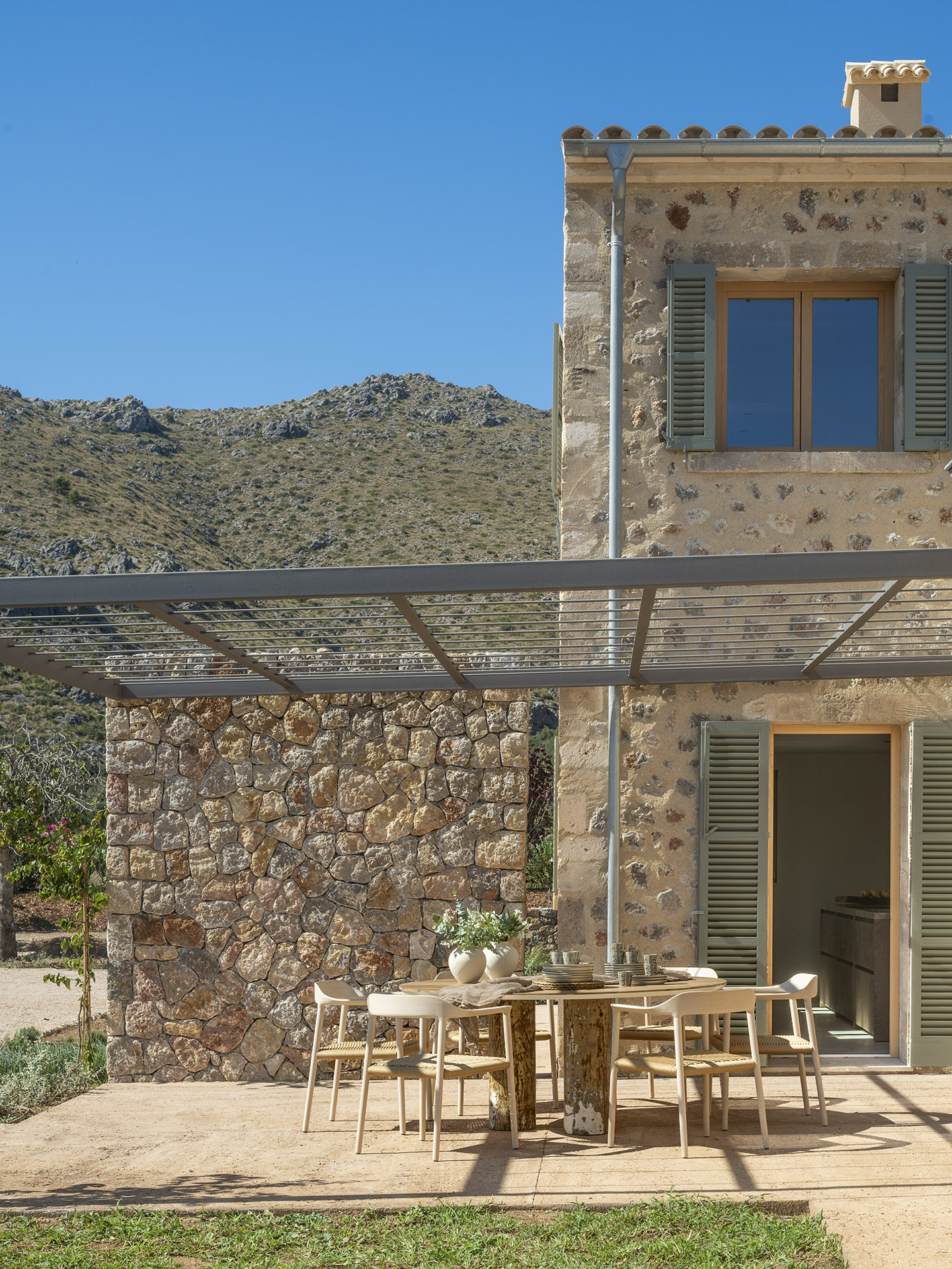 Steinfassade mit grünen Fensterläden, Metallpergola und Außentisch mit hellen Stühlen vor der Bergkulisse