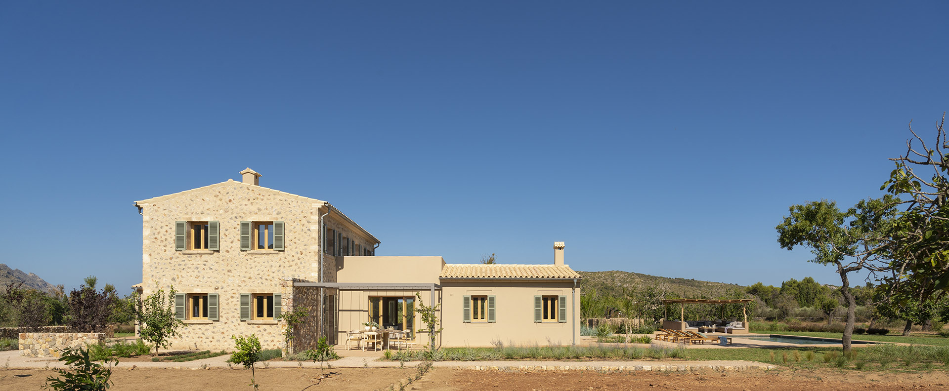 Zweigeschossiges Steinhaus mit eingeschossigem Anbau, blauem Himmel und Swimmingpool im Hintergrund