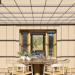 Essbereich im Freien mit rundem Tisch und hellen Stühlen unter einer Metallpergola, neben einem großen Fenster