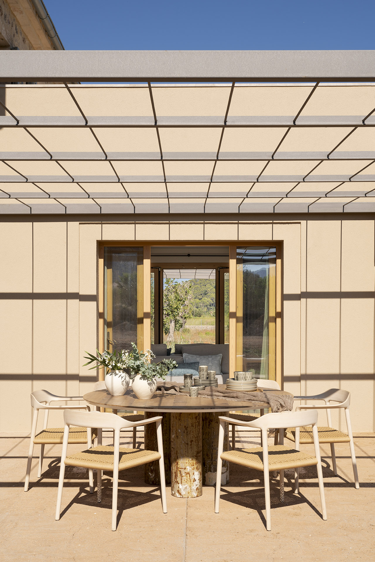 Essbereich im Freien mit rundem Tisch und hellen Stühlen unter einer Metallpergola, neben einem großen Fenster