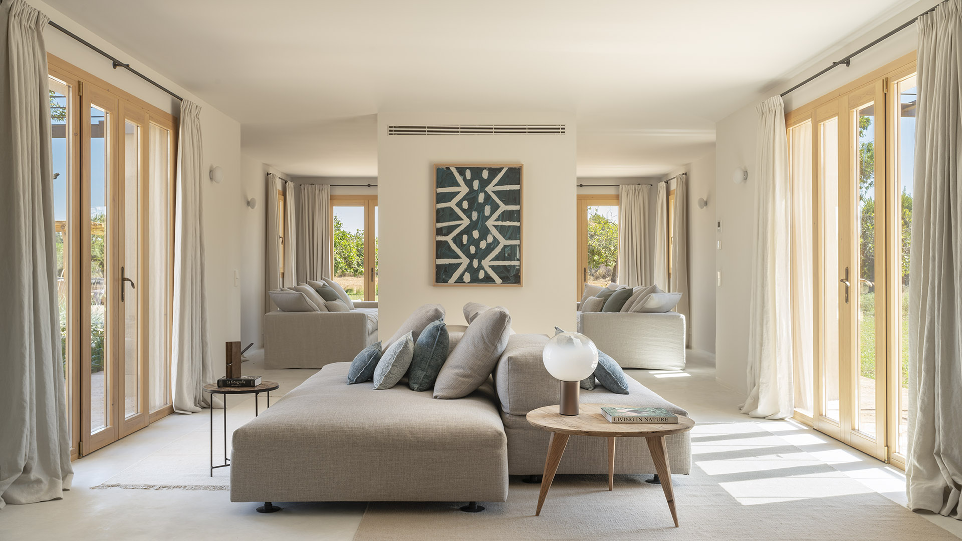 Helles Wohnzimmer mit grauem Modulsofa, blaugrünem Gemälde, langen Vorhängen und großen Holzfenstern zum Garten