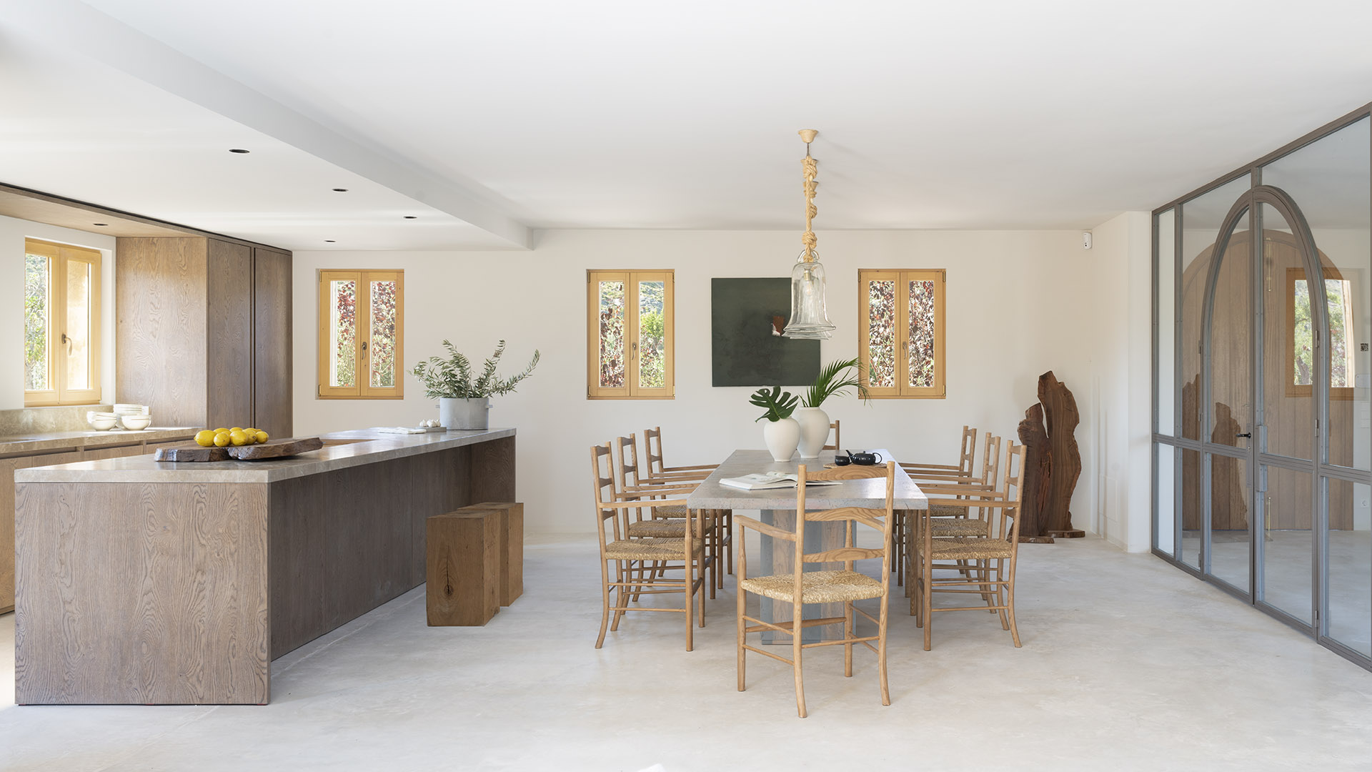 Küche mit Kochinsel aus Holz, offen zum Essbereich, mit langem Tisch, Holzfenstern und einer Glasfront im Rundbogen