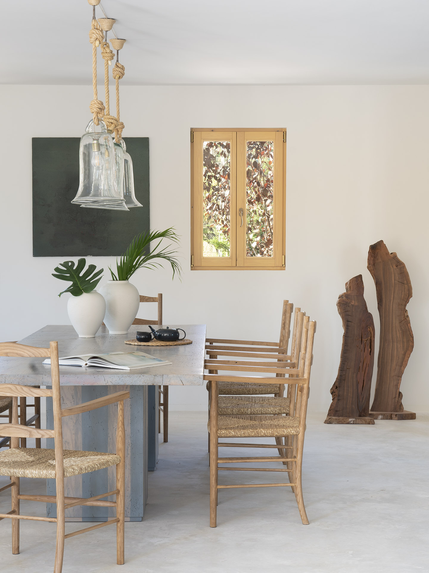 Detailansicht des Essbereichs mit grauem Tisch, Holzstühlen, Glaslampen, grünem Gemälde und skulptural geformten Baumstämmen