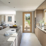 Küche mit Kochinsel und Steinarbeitsplatte, Holzschränken, Gartentür und Wohnzimmer im Hintergrund