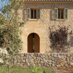 Bogentür aus Holz in einer Natursteinfassade, mit grünen Fensterläden, Olivenbaum und Steinmauer