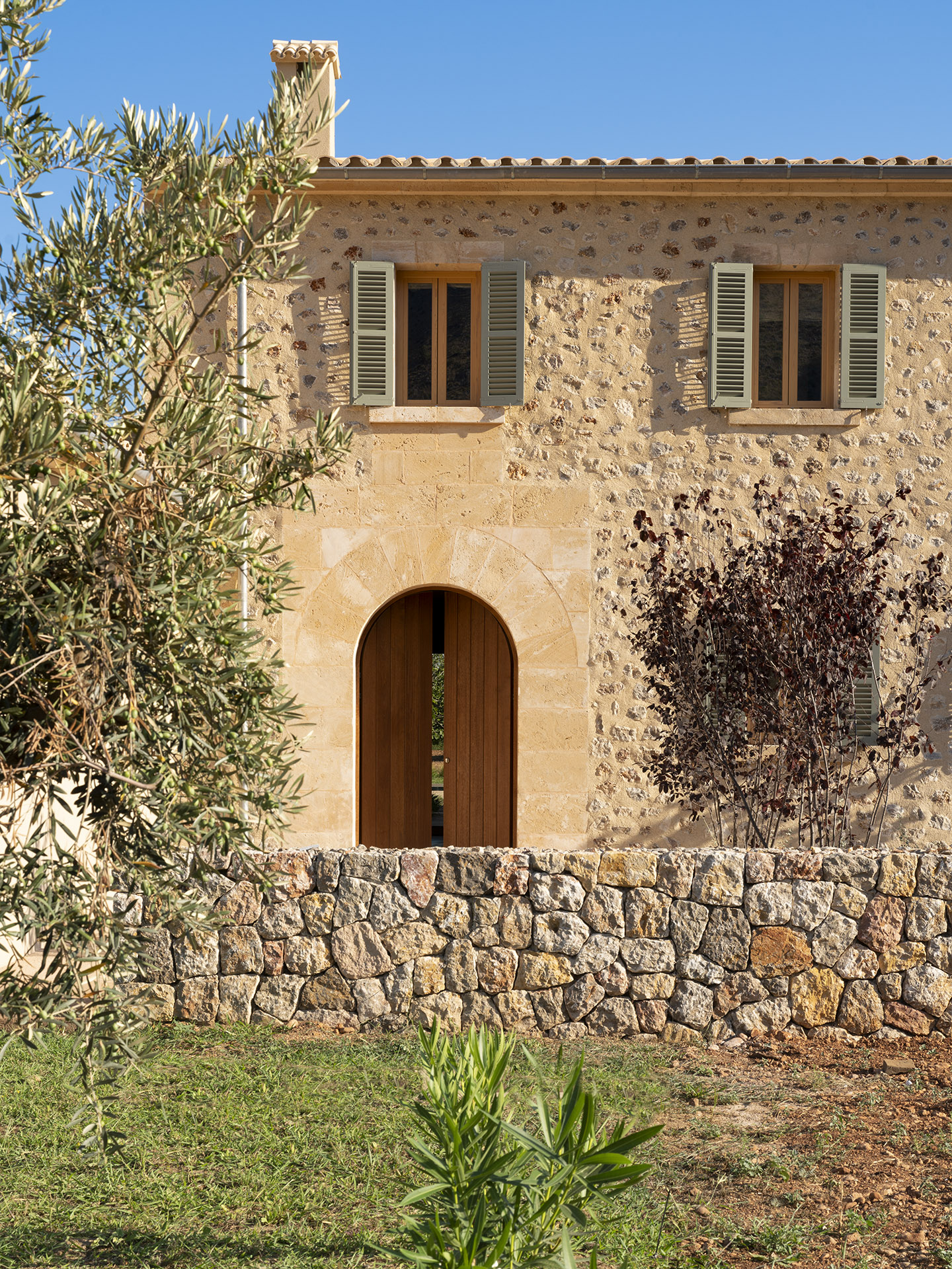 Bogentür aus Holz in einer Natursteinfassade, mit grünen Fensterläden, Olivenbaum und Steinmauer