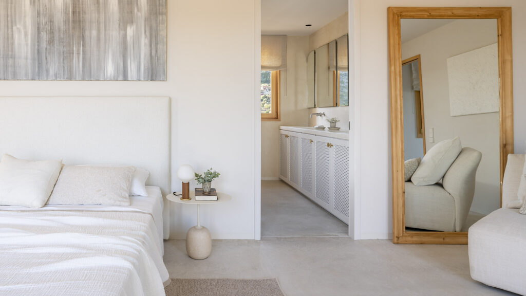 Schlafzimmer in Ecru-Tönen mit weißem Kopfteil, Holzspiegel und Ensuite-Bad im Hintergrund