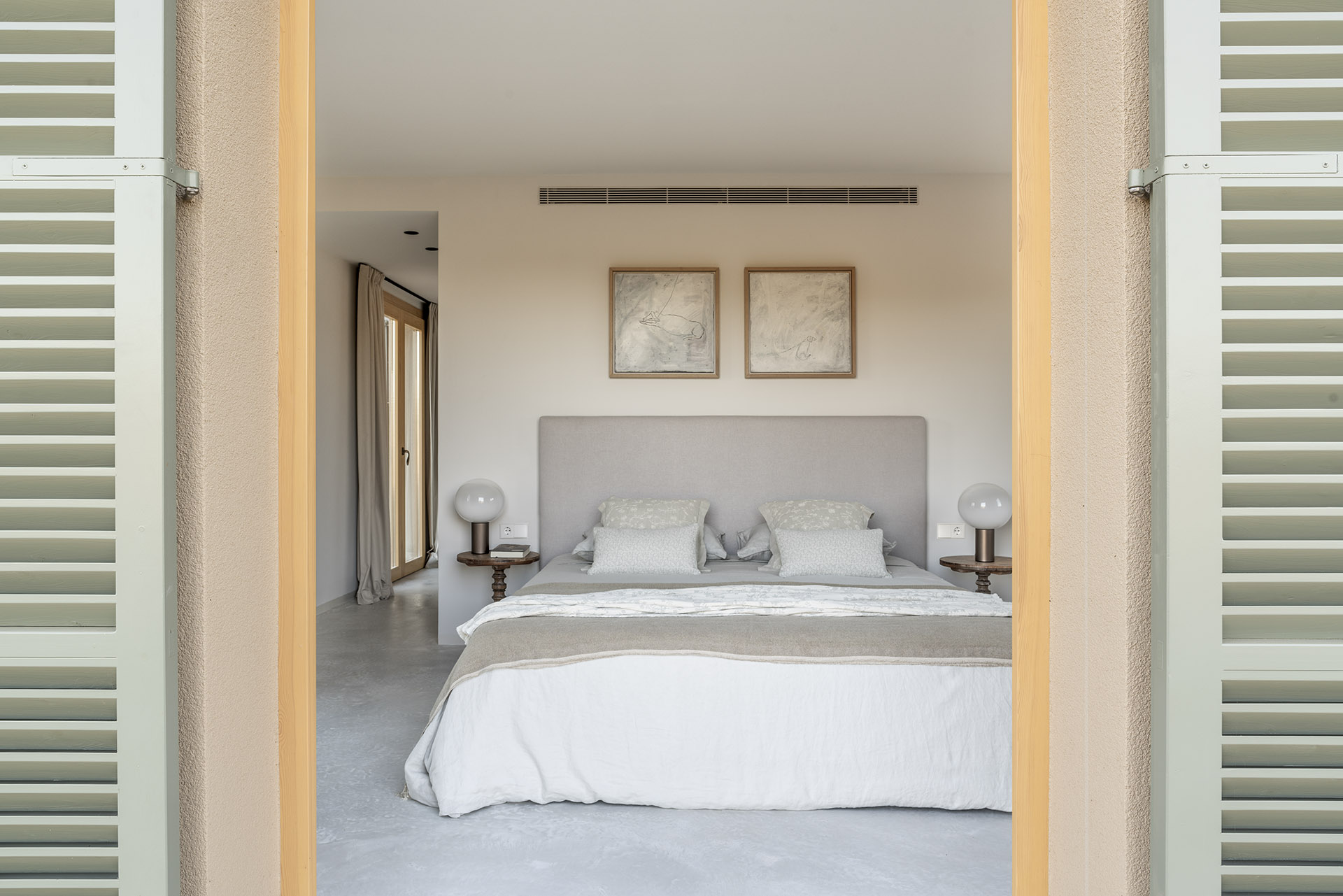 Schlafzimmer zwischen offenen mallorquinischen Fensterläden, mit grauem Kopfteil, zwei Bildern und poliertem Betonboden