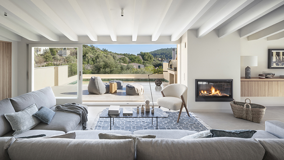 Salón de SJ61 con chimenea encendida, vigas blancas y terraza con piscina al fondo
