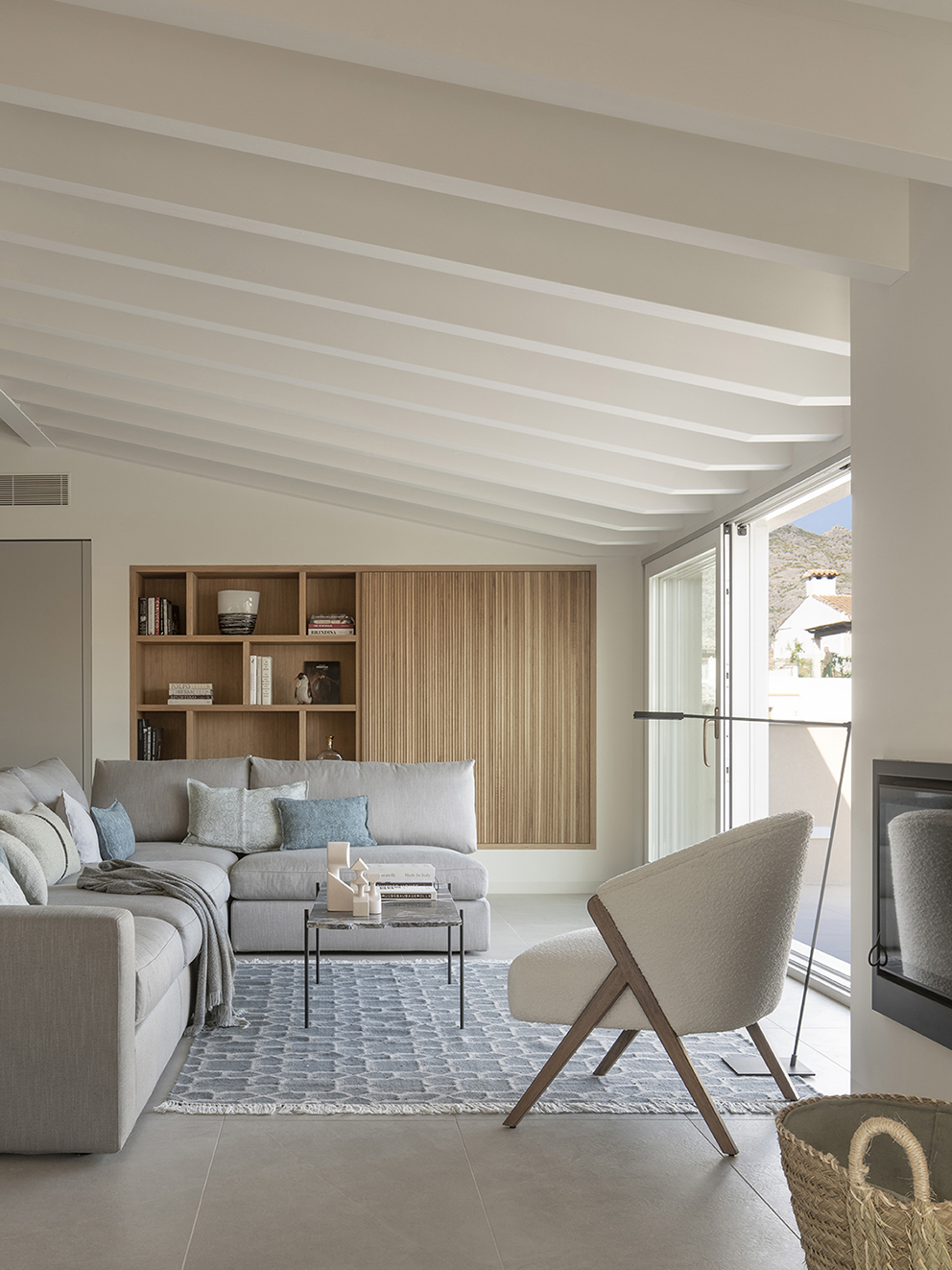 Corner of the living room with light wood shelving, grey sofa, white armchair and beamed ceiling