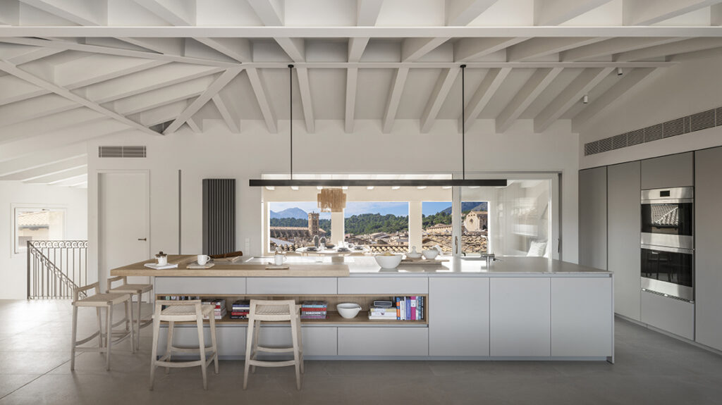 Kitchen with a large white island, light wood breakfast bar, stools and a window overlooking the village