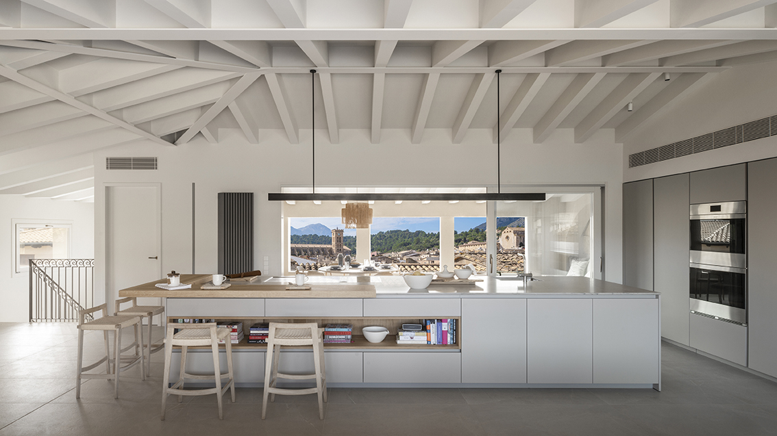 Kitchen with a large white island, light wood breakfast bar, stools and a window overlooking the village