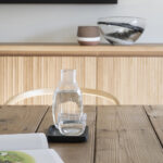 Detail of a jug and glass of water on a rustic wooden table, with a fluted sideboard behind