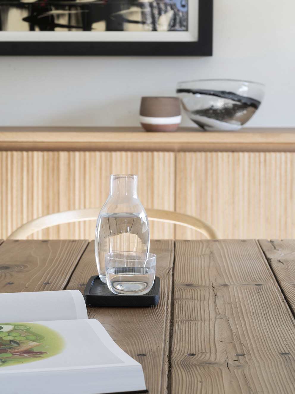 Detail of a jug and glass of water on a rustic wooden table, with a fluted sideboard behind
