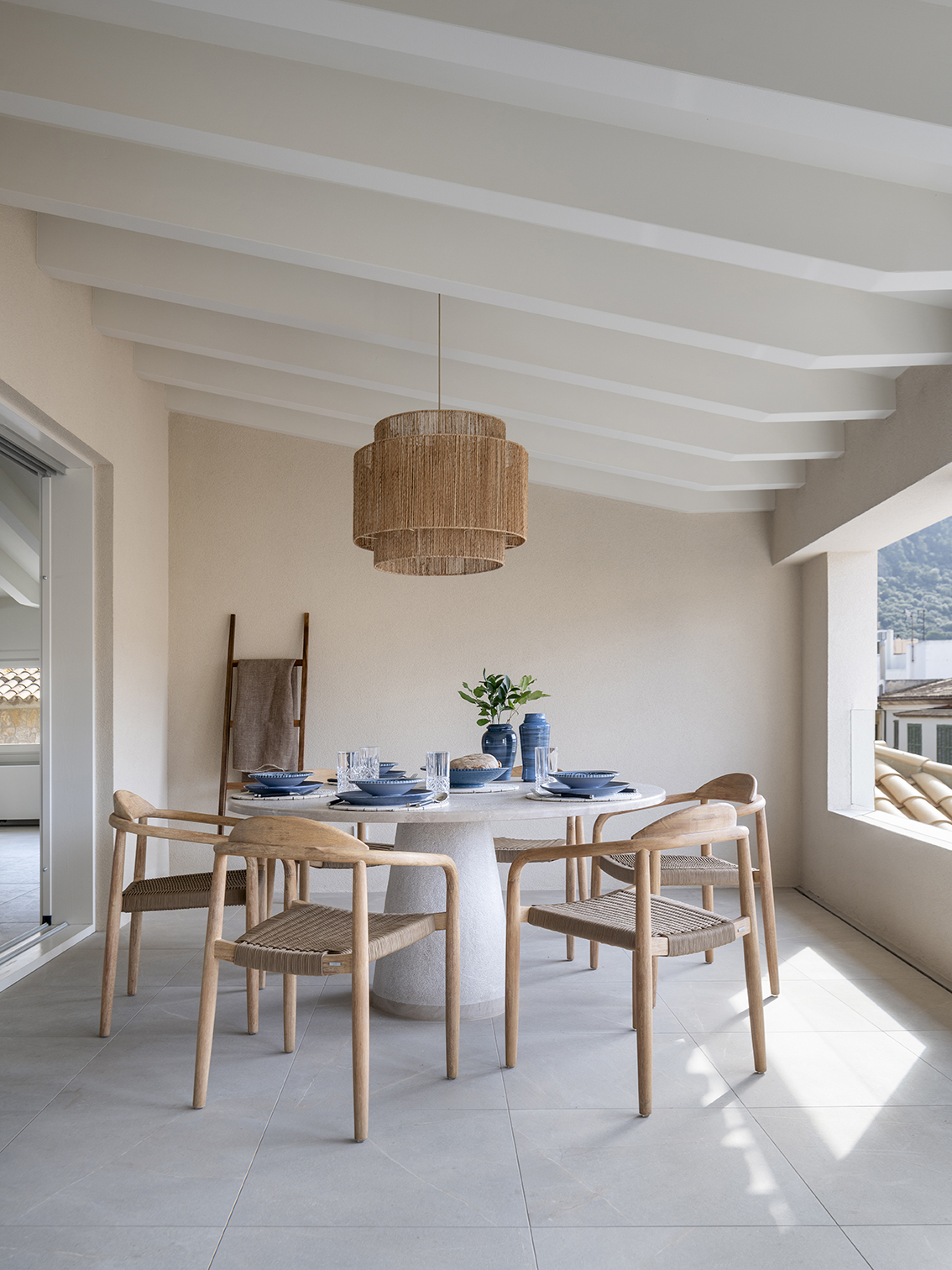 Terrace with a round white table, blue tableware, wooden chairs and a natural fibre lamp