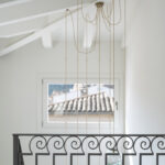 Pendant lamps with rope cord beside a wrought iron balustrade and a window overlooking tiled roofs