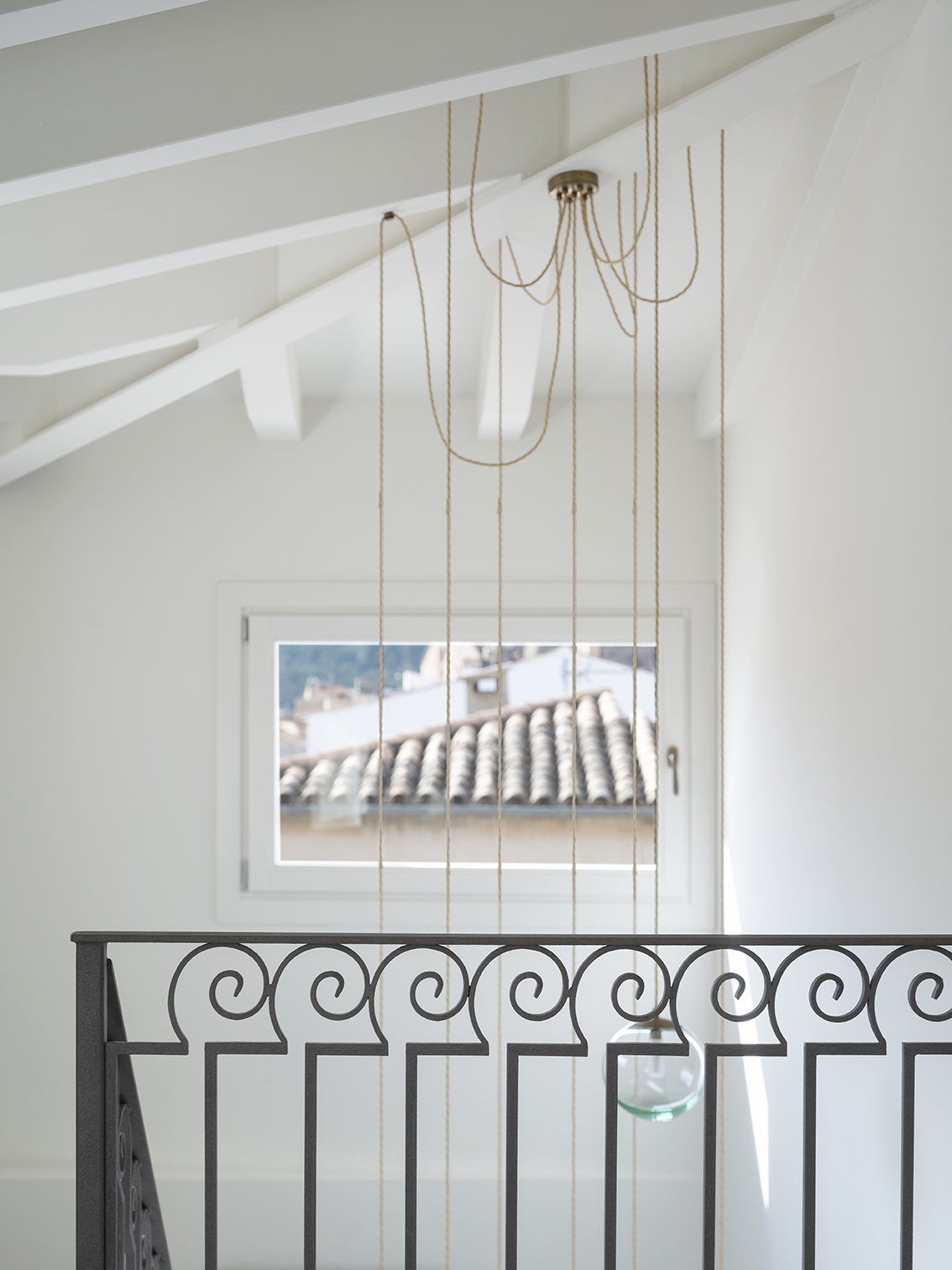 Pendant lamps with rope cord beside a wrought iron balustrade and a window overlooking tiled roofs