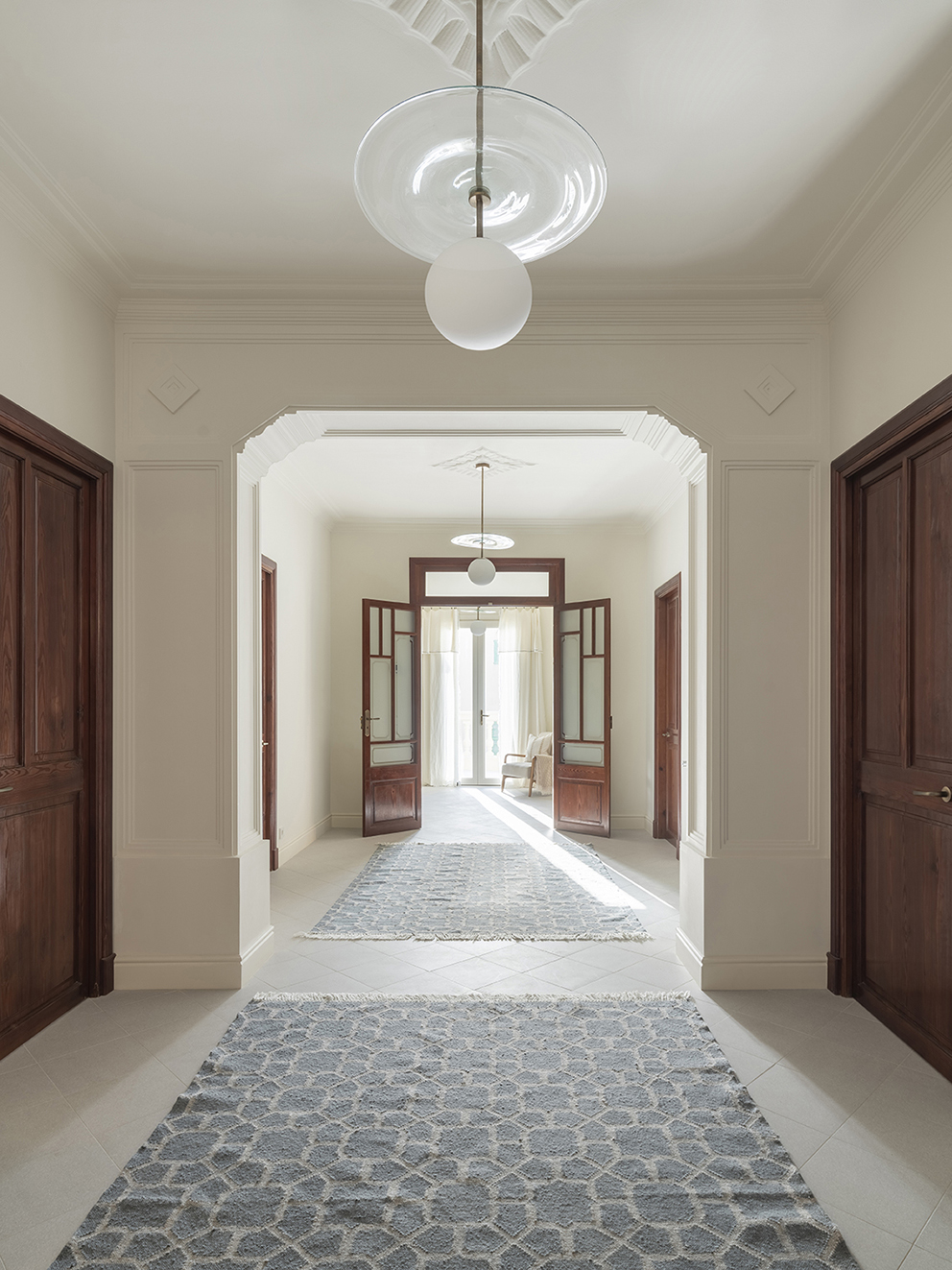 Wide hallway with moulded arch, dark wooden doors, blue-grey rugs and natural light at the far end
