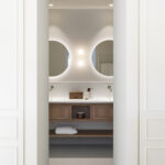 Bathroom with two round backlit mirrors and a wooden unit, seen between white wardrobes