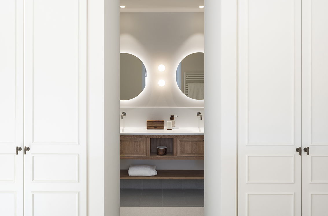 Bathroom with two round backlit mirrors and a wooden unit, seen between white wardrobes