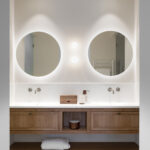 Front view of the double washbasin with wooden unit, white worktop and round backlit mirrors