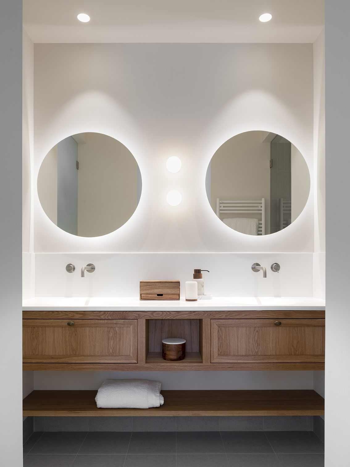 Front view of the double washbasin with wooden unit, white worktop and round backlit mirrors
