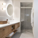 Bathroom with wooden vanity unit, round backlit mirror and glass shower with vertical tiles