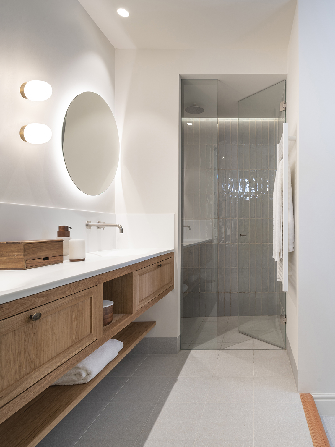 Bathroom with wooden vanity unit, round backlit mirror and glass shower with vertical tiles