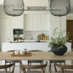 White kitchen island behind the dining table, with pendant lights and a black vase of branches
