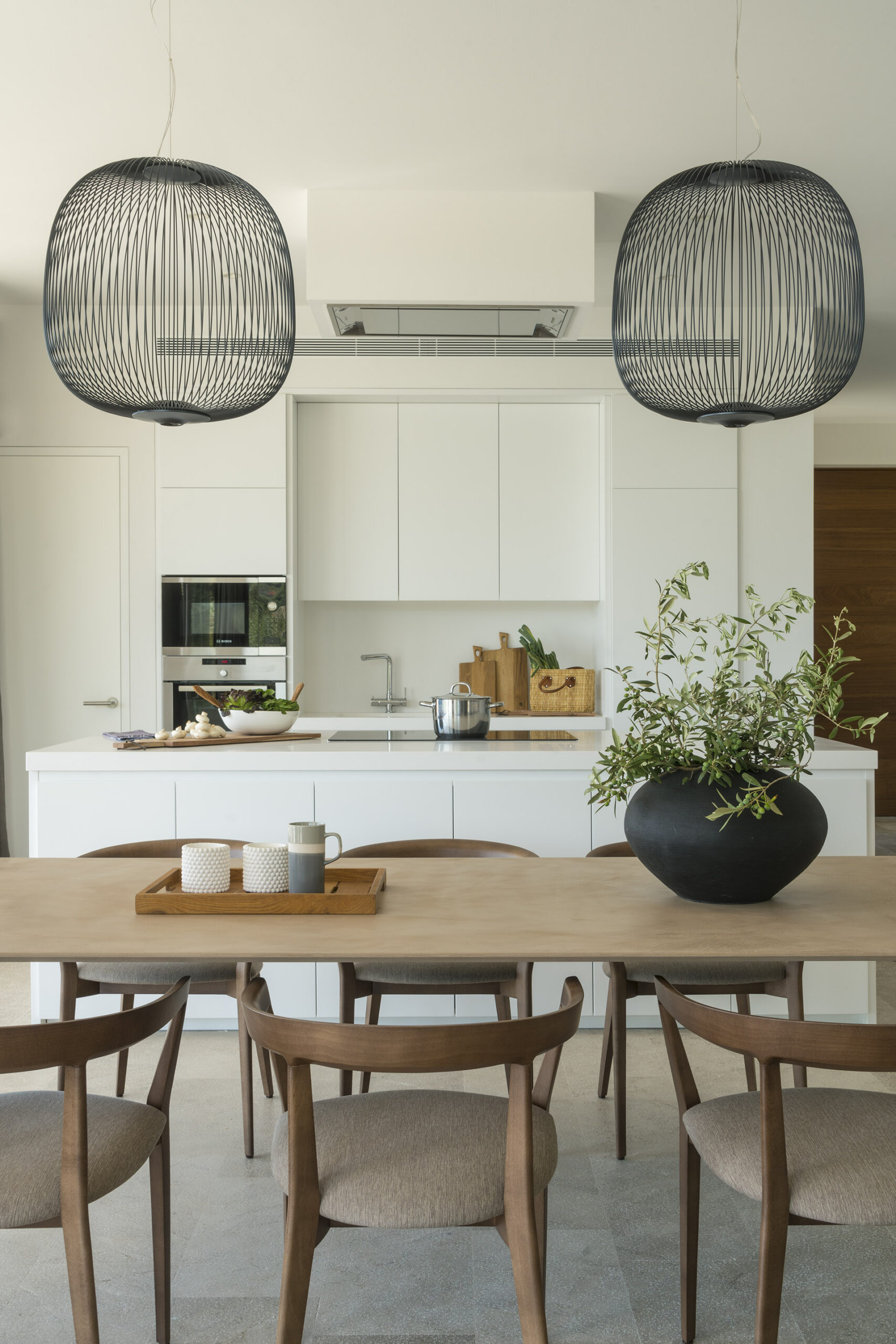 White kitchen island behind the dining table, with pendant lights and a black vase of branches