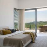 Bedroom with beige bedspread, mustard throw and sliding door to a terrace with views of the hills
