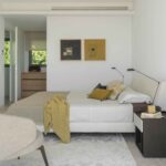 Bedroom with upholstered headboard, two abstract paintings, wooden chest of drawers at the back and stone floor