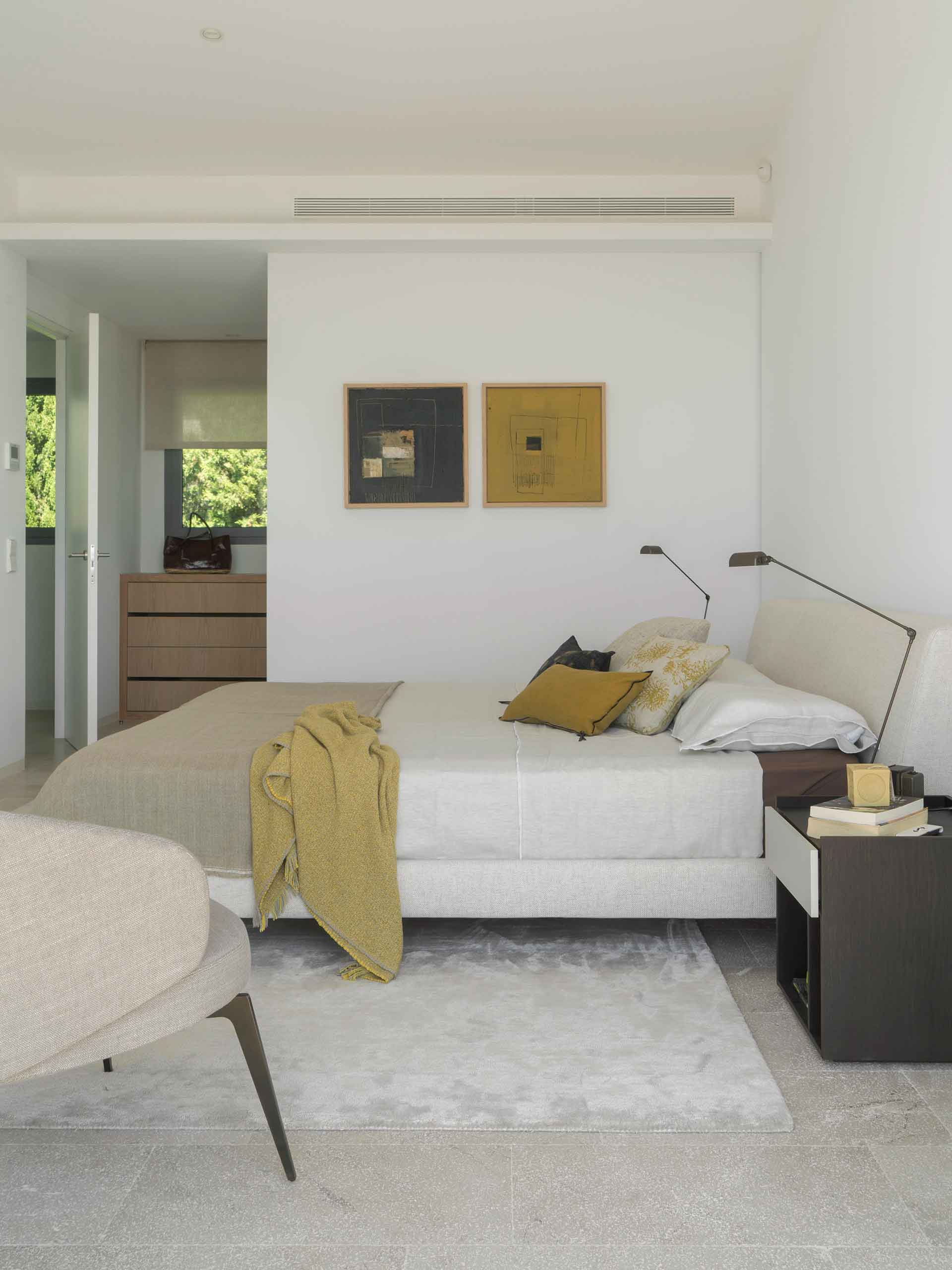Bedroom with upholstered headboard, two abstract paintings, wooden chest of drawers at the back and stone floor