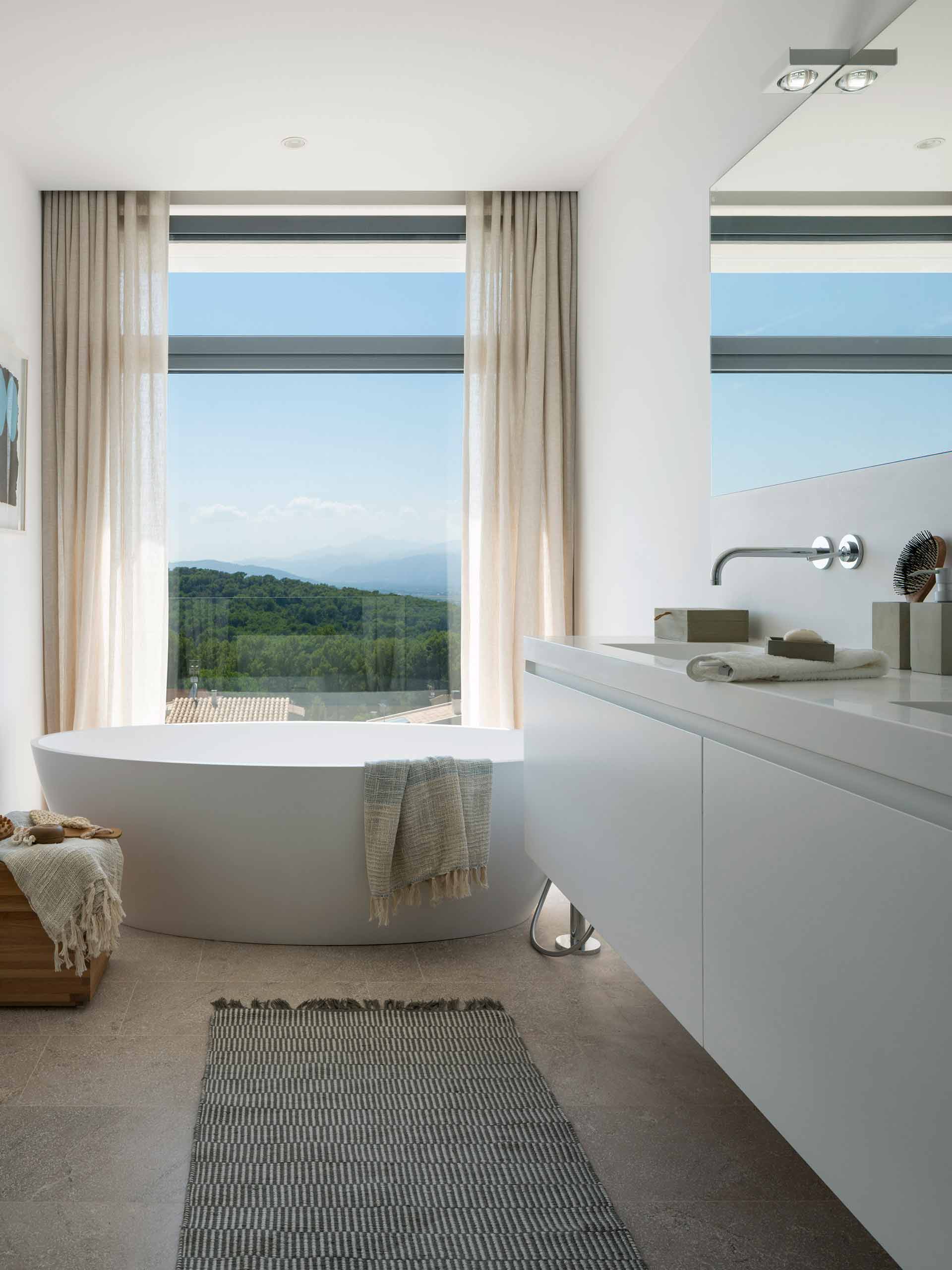 Bathroom with oval freestanding bath in front of a large window with hill views and wall-hung basin