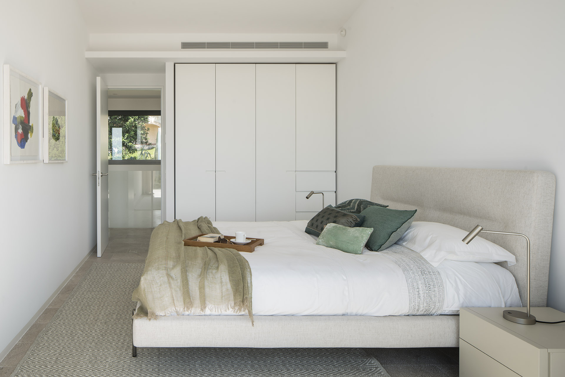 Bedroom with white built-in wardrobe, bed with beige headboard, green cushions and colourful paintings