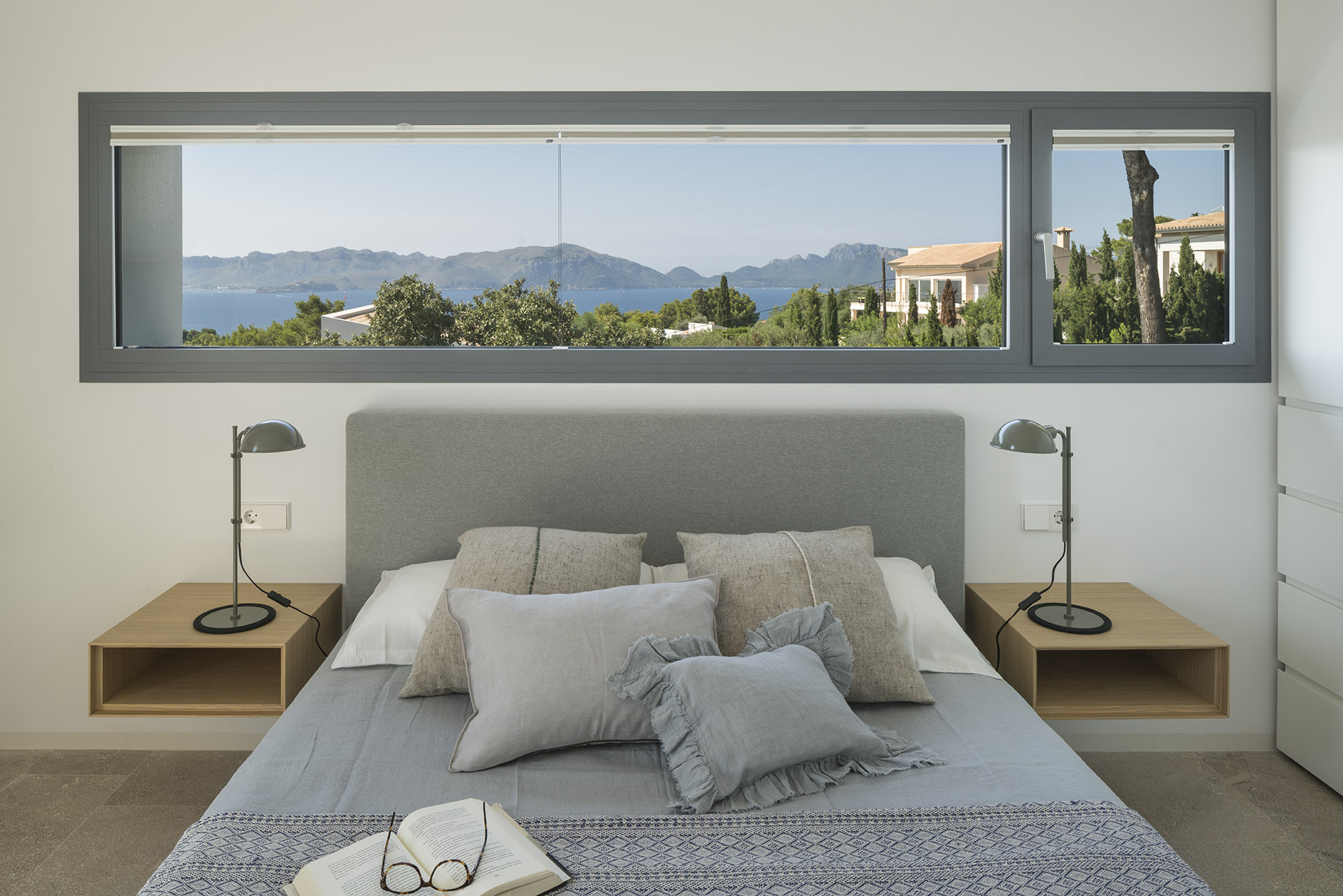 Grey headboard between two floating wooden bedside tables, with a horizontal window overlooking the sea