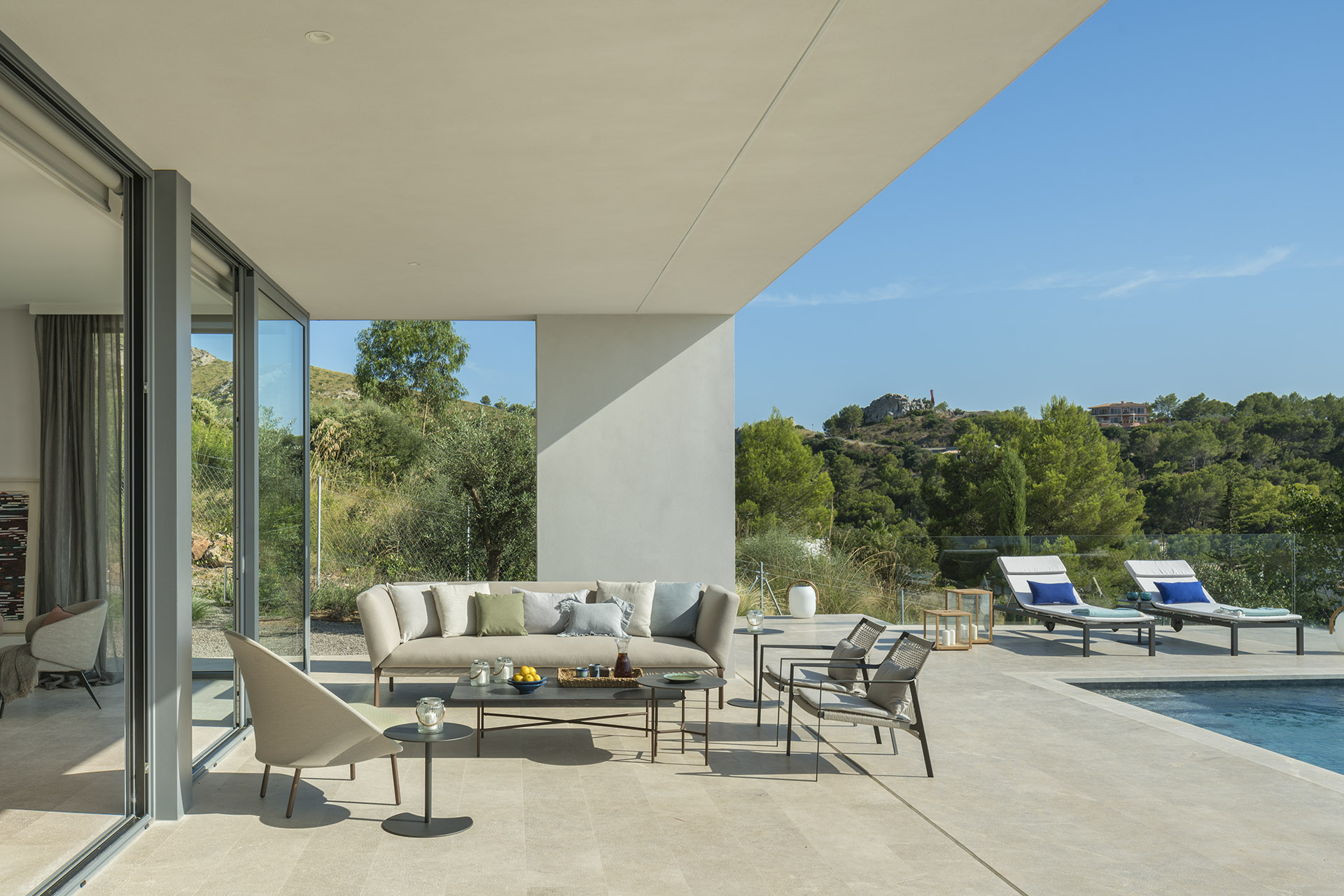 Covered terrace with beige sofa, armchair, large glass windows onto the living room and pool on the right