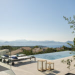 View of the infinity pool with sun loungers, lanterns and an olive tree, and the sea with mountains beyond