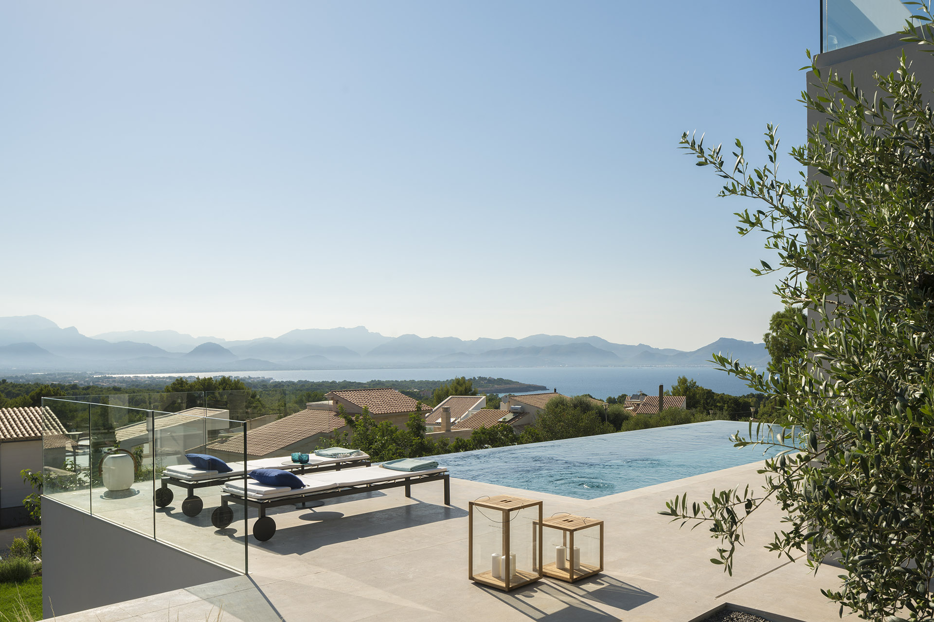 View of the infinity pool with sun loungers, lanterns and an olive tree, and the sea with mountains beyond